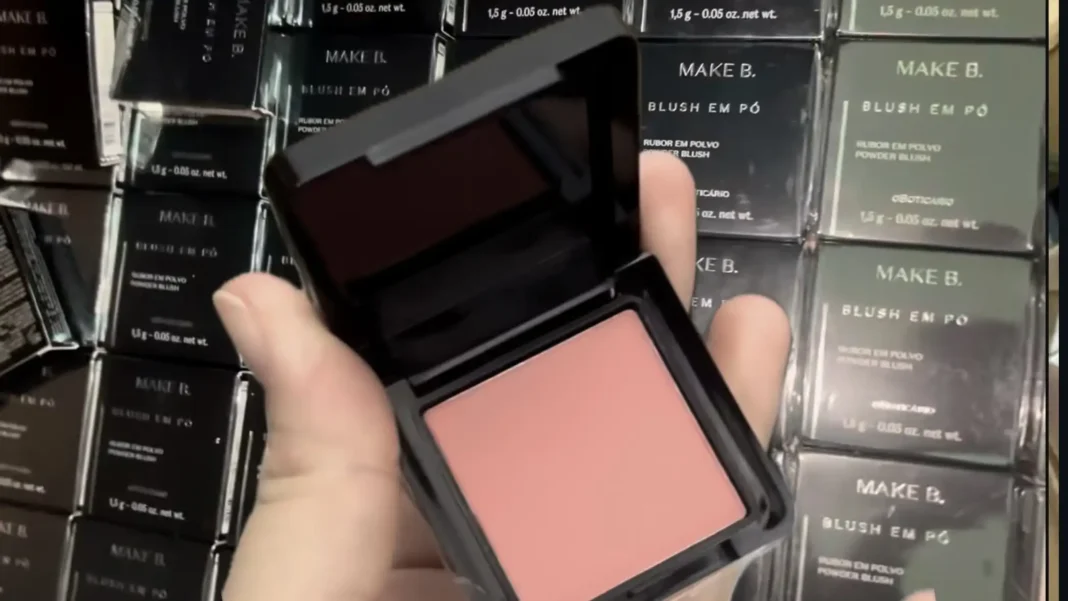 Uma mão segura um estojo quadrado preto de maquiagem Make B. aberto, revelando um blush em pó cor de pêssego. Caixas do produto empilhadas são visíveis ao fundo.