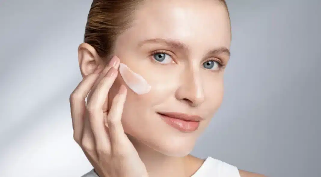 Uma mulher sorridente com cabelos loiros e olhos azuis está aplicando um produto de cuidados com a pele em sua bochecha. O produto é uma loção cremosa e branca, e ela o aplica suavemente com a ponta dos dedos. A imagem tem um fundo cinza e foca em seu rosto e no produto.