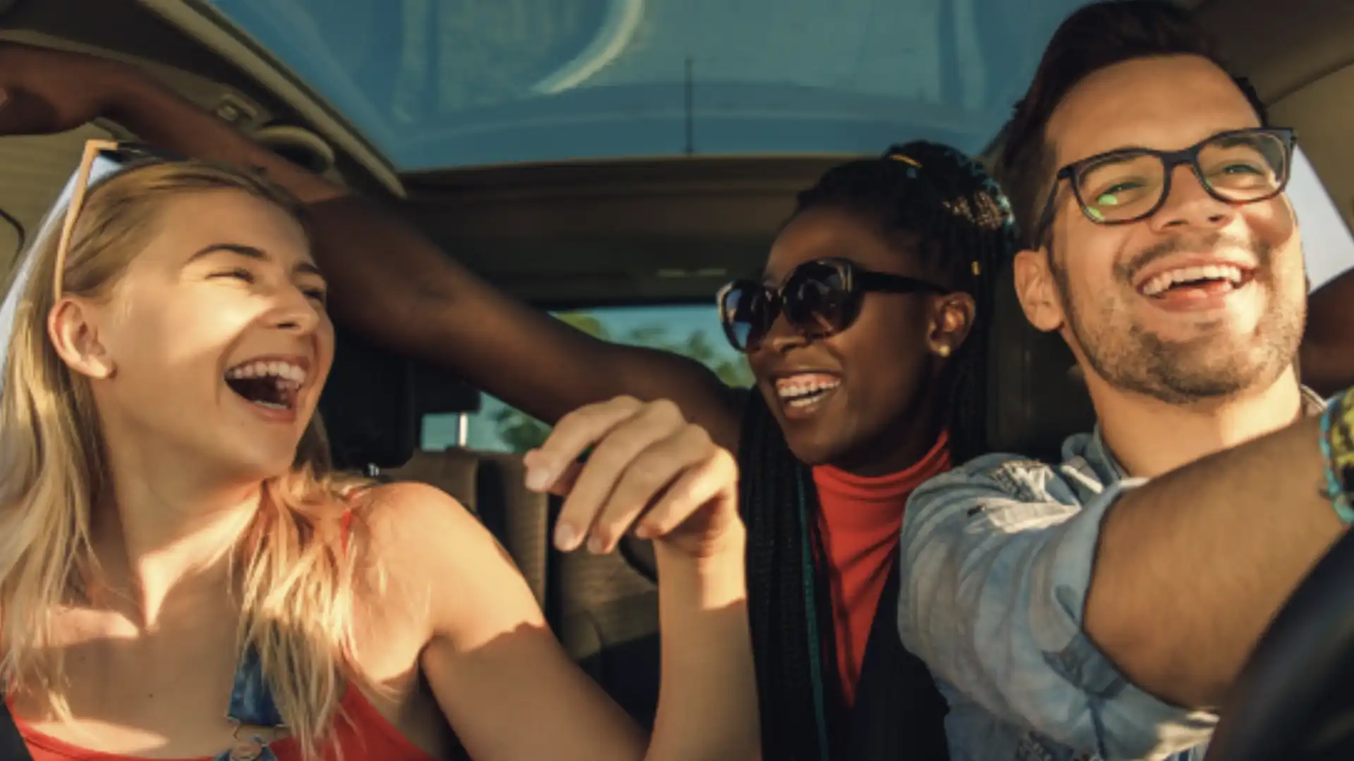 Três amigos rindo dentro de um carro com o teto solar aberto. O homem que está dirigindo usa óculos e sorri amplamente. As duas mulheres ao lado também sorriem.