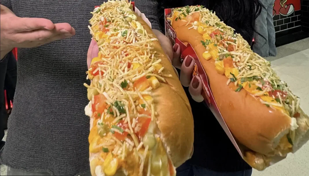 Duas pessoas segurando cachorros-quentes enormes e recheados, um ao lado do outro, em uma embalagem de papelão.