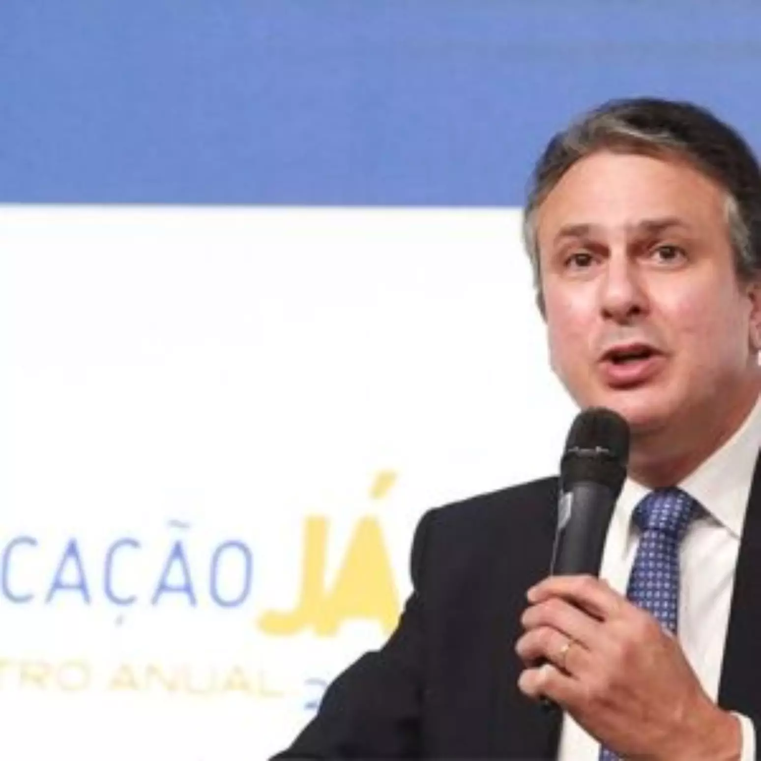 Imagem em close-up de um homem de meia-idade, com cabelos castanhos e curtos, vestindo terno azul escuro e gravata com pontos brancos, falando em um microfone.