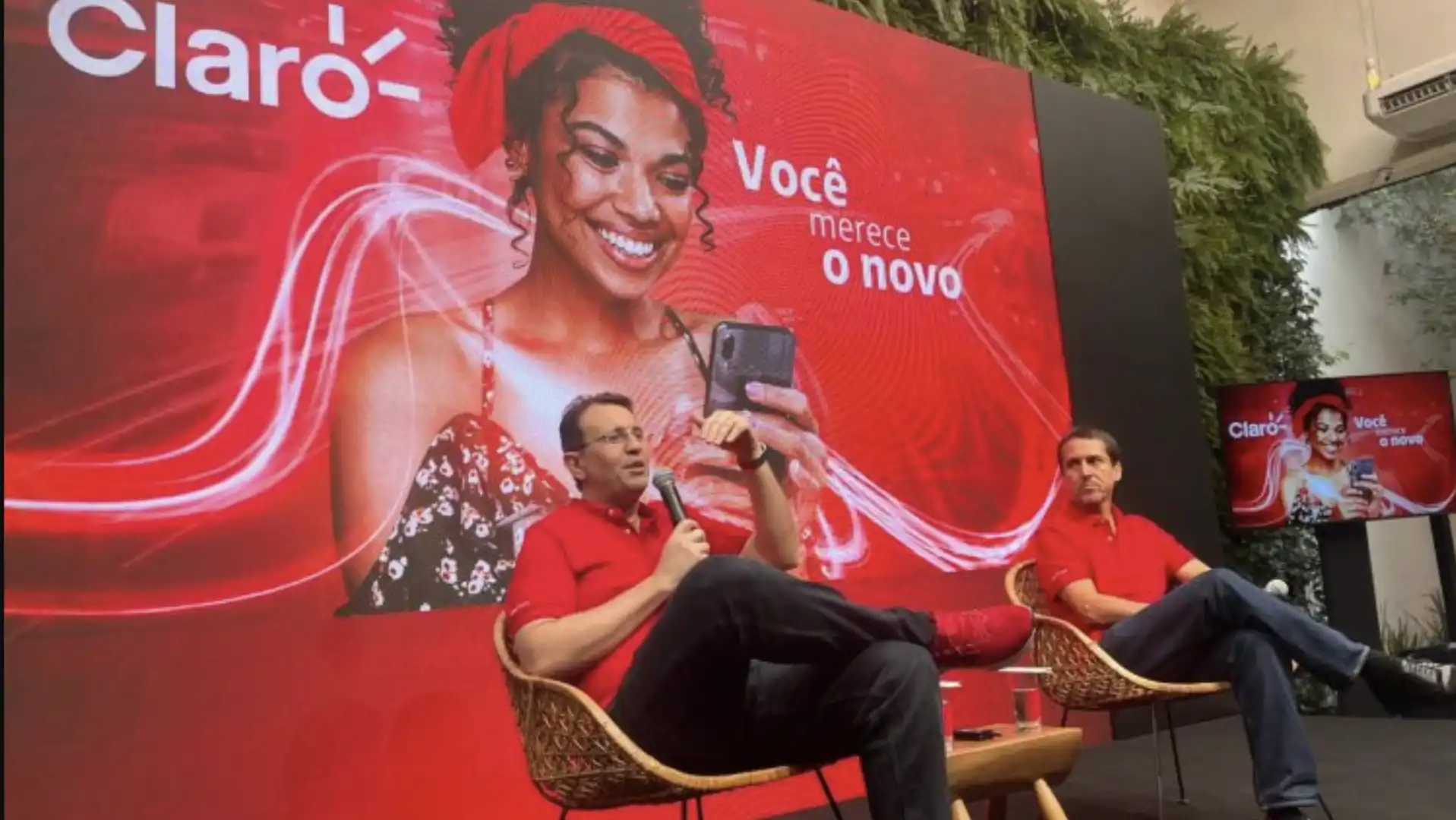 Dois homens em poltronas, um deles segurando um celular, com um painel grande da Claro ao fundo.