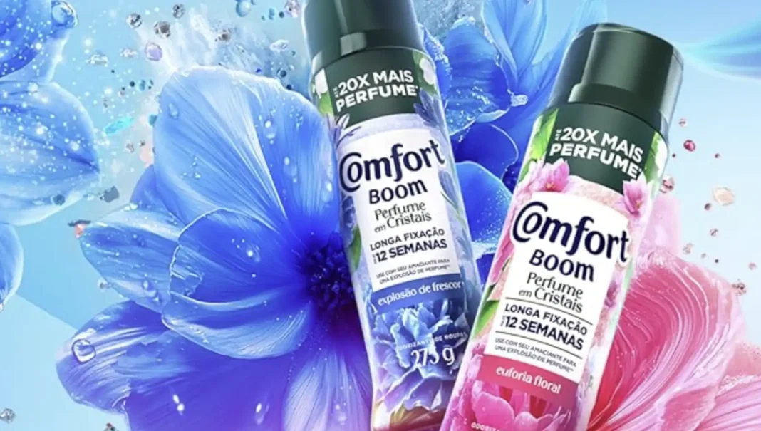 Duas garrafas de amaciante Comfort Boom de perfume em cristais, uma azul e uma rosa, flutuando em um fundo de flores azuis e rosas com brilhos.