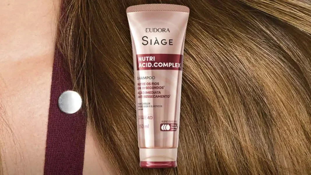 Mulher com cabelos loiros esvoaçantes e ondulados, olhando para a câmera. Ao lado dela está um tubo de shampoo Eudora Siàge Nutri.Acid.Complex.