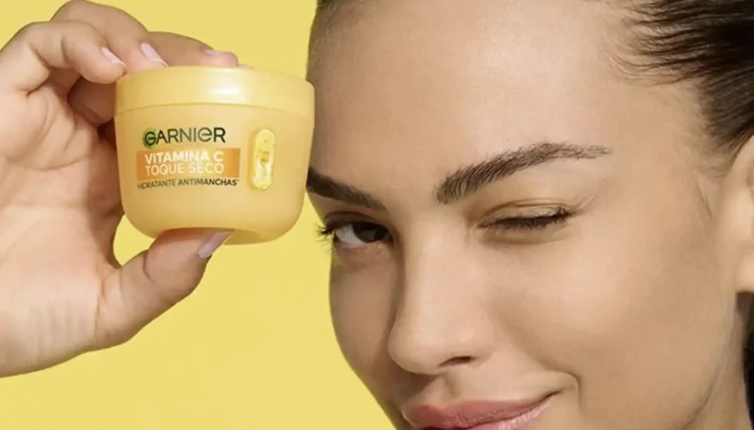 Uma mulher sorridente e com o olho direito piscando segura um pote amarelo de um creme facial da Garnier. No pote está escrito 