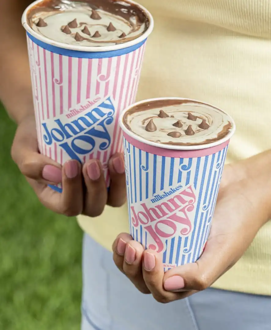 Uma pessoa segurando dois milkshakes da marca "Johnny Joy", um grande e um pequeno. Os milkshakes têm cobertura de chocolate e pequenos pedaços de chocolate.