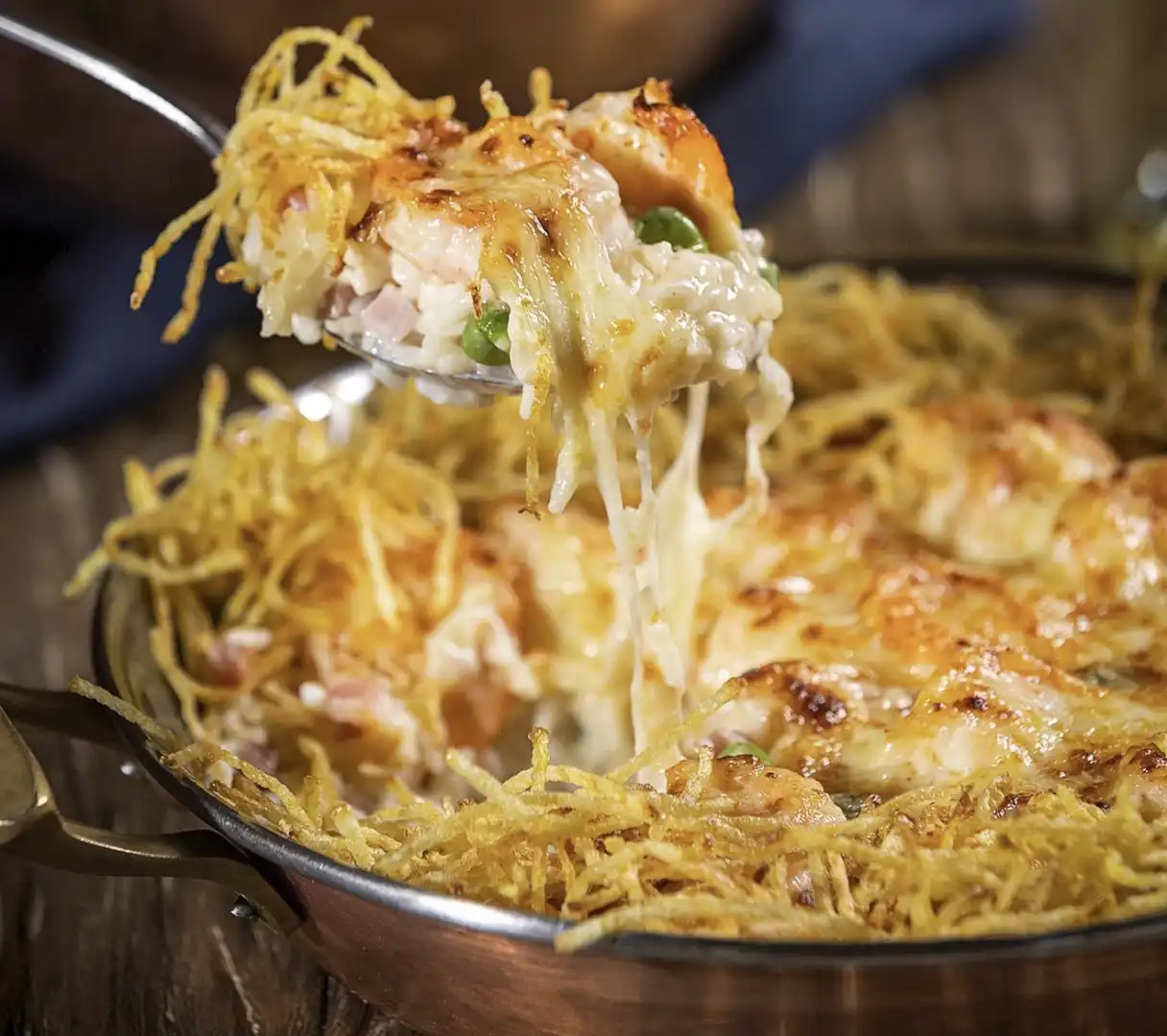 Uma colher cheia de camarão cremoso e gratinado com queijo sendo retirada de uma panela cheia de batata palha.