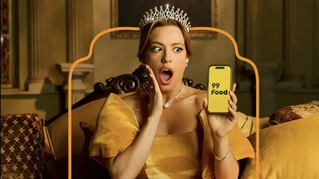 Mulher chocada com coroa e vestido amarelo, segurando celular com logo 