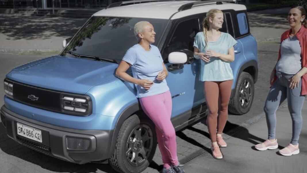 Três mulheres em roupas esportivas conversam ao lado de um carro compacto SUV azul e branco da Chevrolet, aparentemente em um estacionamento.