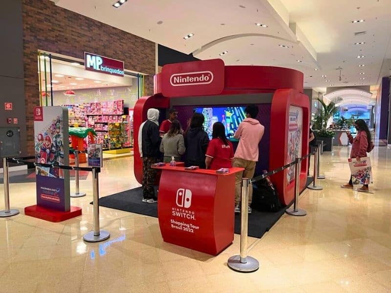 Estande de jogos Nintendo Switch em um shopping, com pessoas jogando e a loja MP Brinquedos ao fundo.