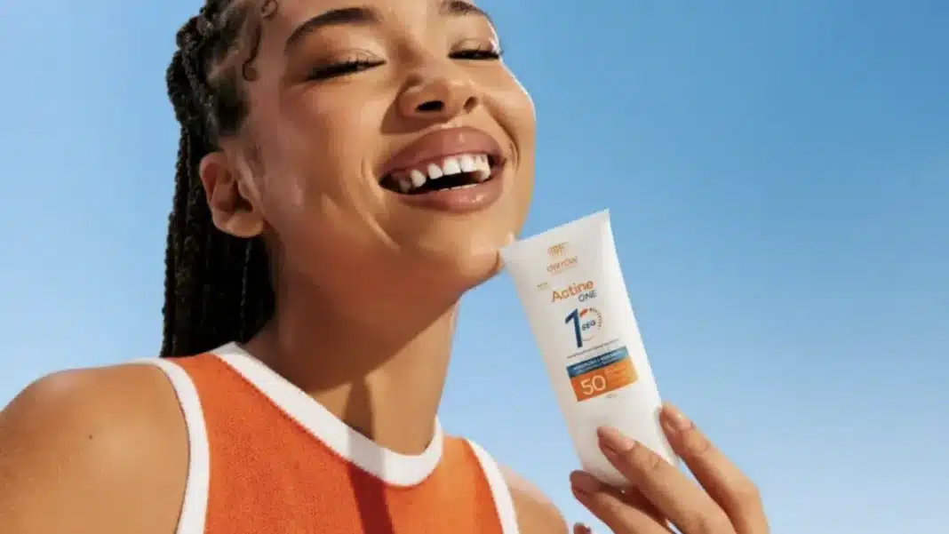 Uma mulher sorridente com tranças, usando um top laranja, segura um tubo de protetor solar branco da marca Actine One. O produto tem um rótulo com a inscrição '1 SEC' e 'FPS 50'.