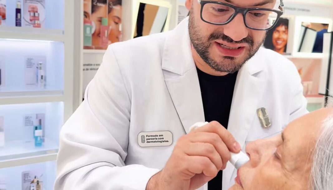 Homem com óculos e jaleco branco aplicando produto de skincare no rosto de uma pessoa mais velha.