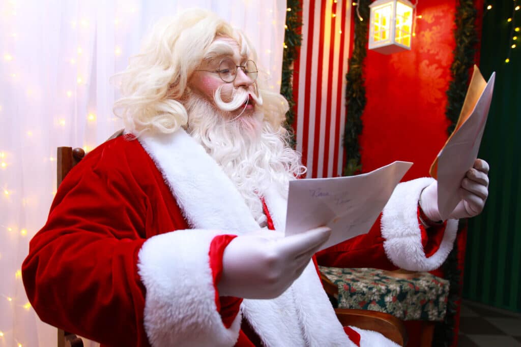 Papai Noel de óculos, barba e cabelos loiros e roupa vermelha, sentado lendo uma carta.