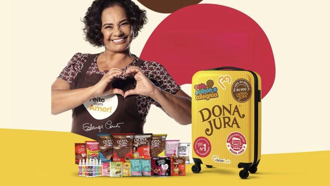 Mulher sorridente da Dona Jura fazendo um coração com as mãos, ao lado de uma mala de viagem amarela com a marca Dona Jura e vários produtos de chocolate na frente.