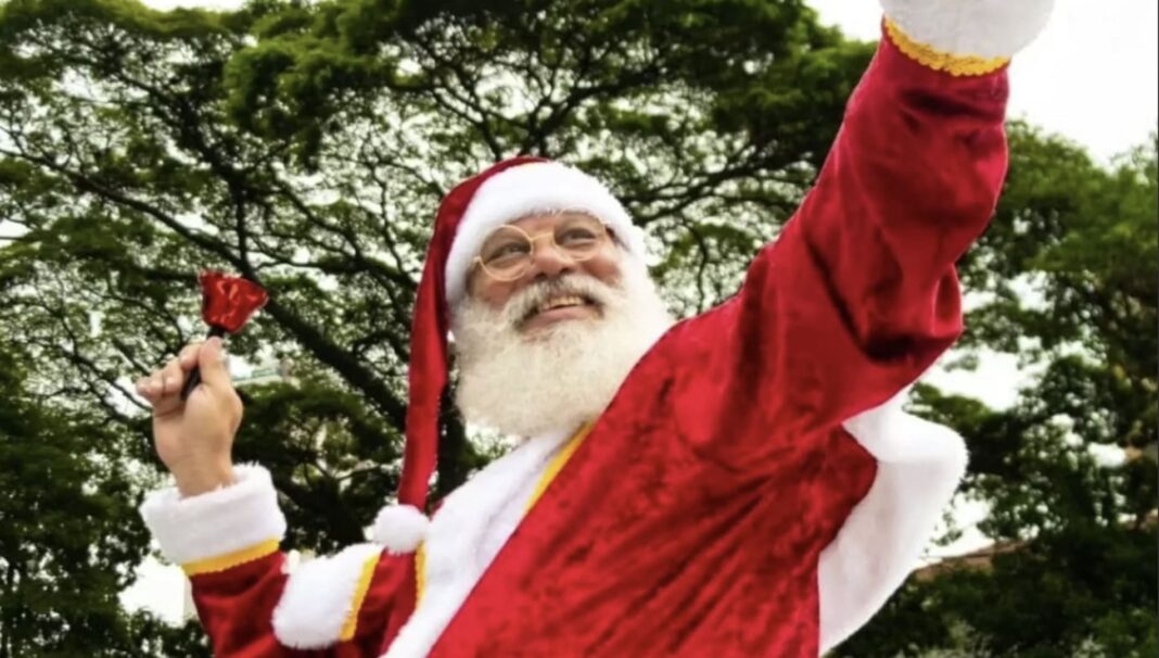Papai Noel sorrindo e acenando, segurando um pequeno sino vermelho.